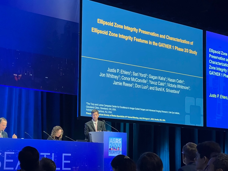 A person standing at a podium with a large screen behind him  Description automatically generated