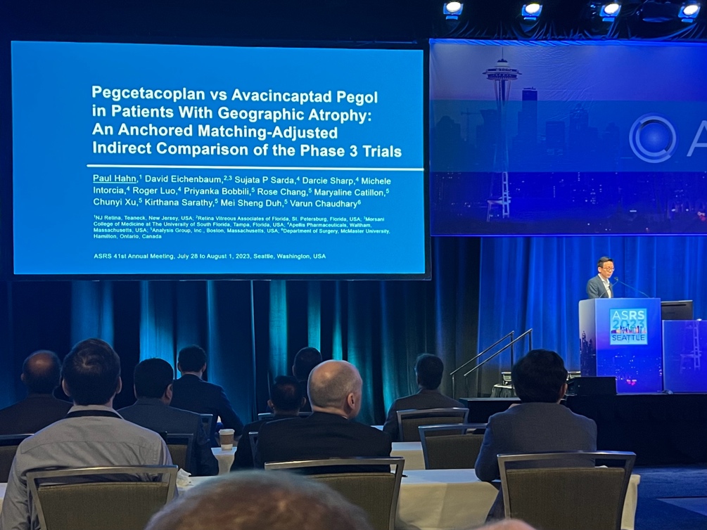 A person standing at a podium with a large screen  Description automatically generated