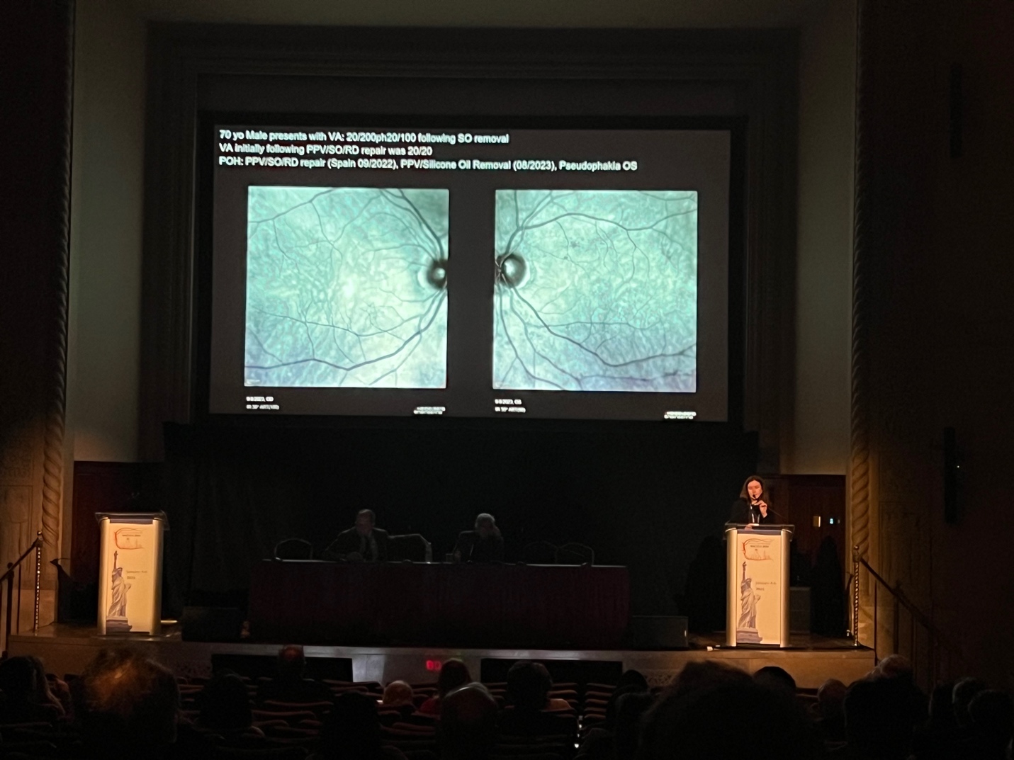 A person standing at a podium with a large screen Description automatically generated